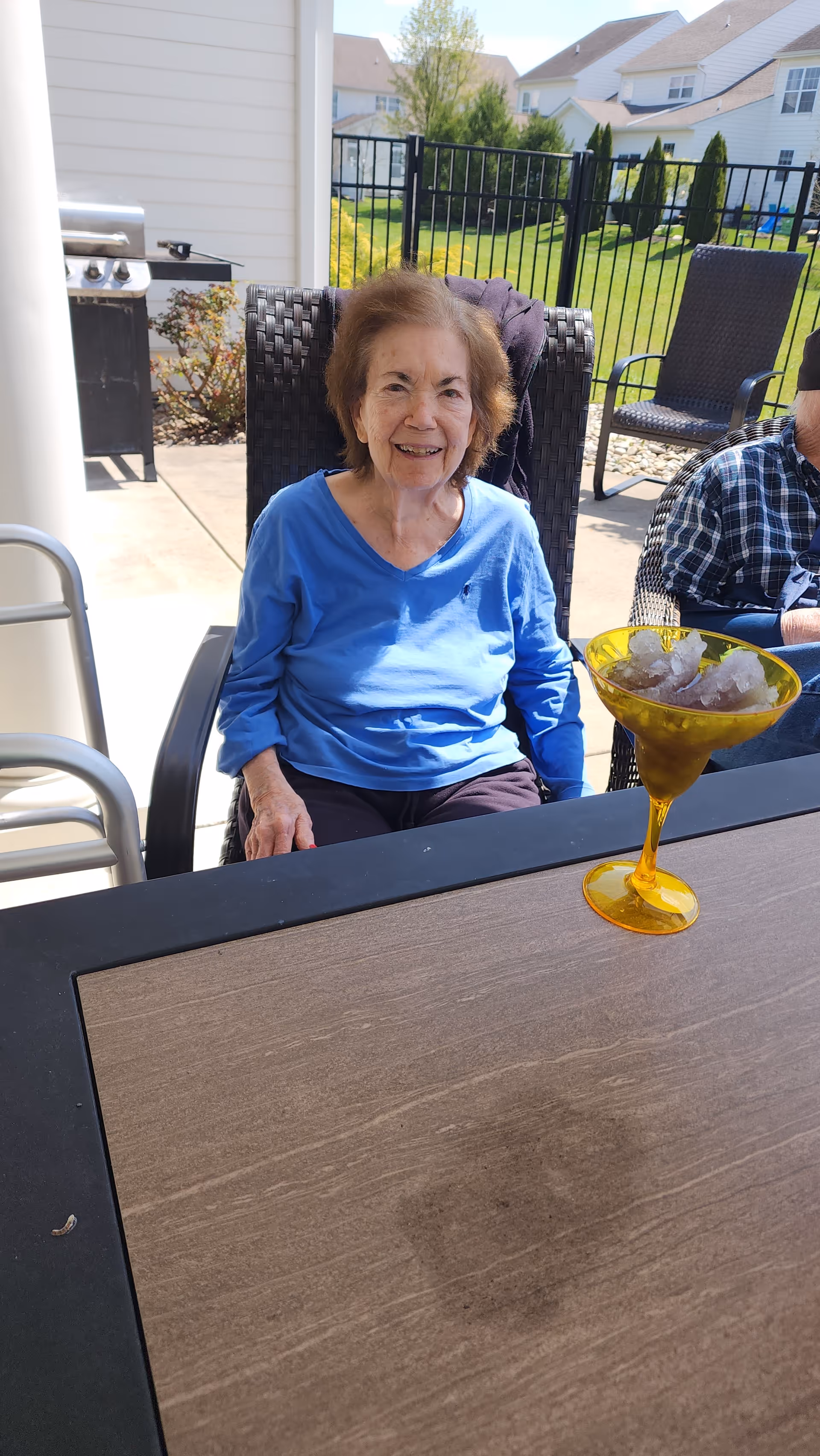 An elderly woman wearing a blue long-sleeve shirt sitting outdoors on a patio chair at a table with a yellow glass filled with ice in front of her. Behind her is a black metal fence, some greenery, and neighboring houses under a clear sky.