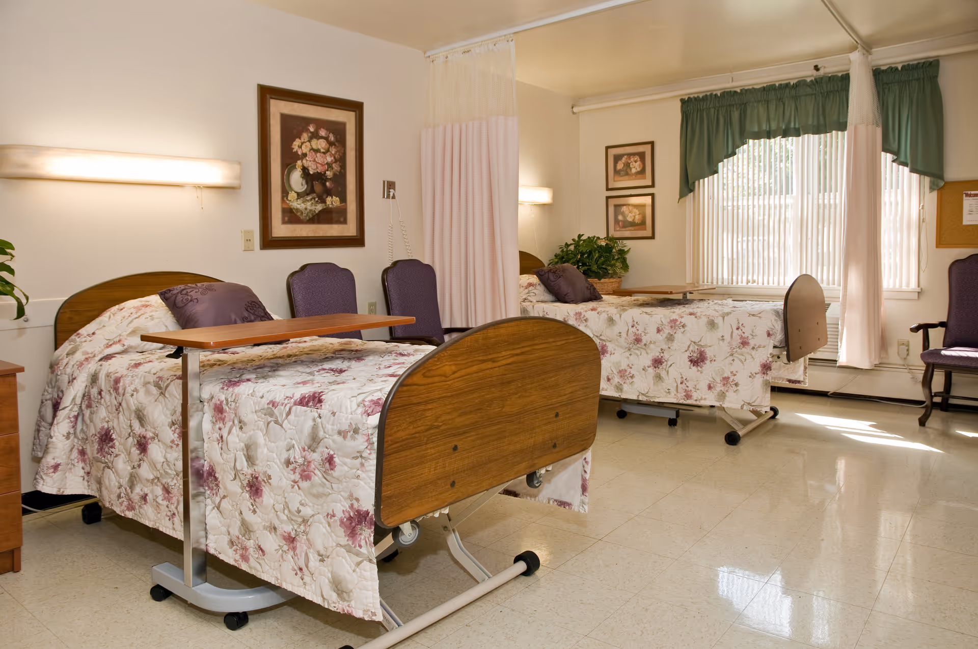 A well-lit room with two hospital-style beds covered in floral bedspreads, each with a purple pillow. There are two purple chairs between the beds and a wooden bedside table. The room has a large window with vertical blinds and green valances, floral paintings on the walls, and a privacy curtain between the beds.