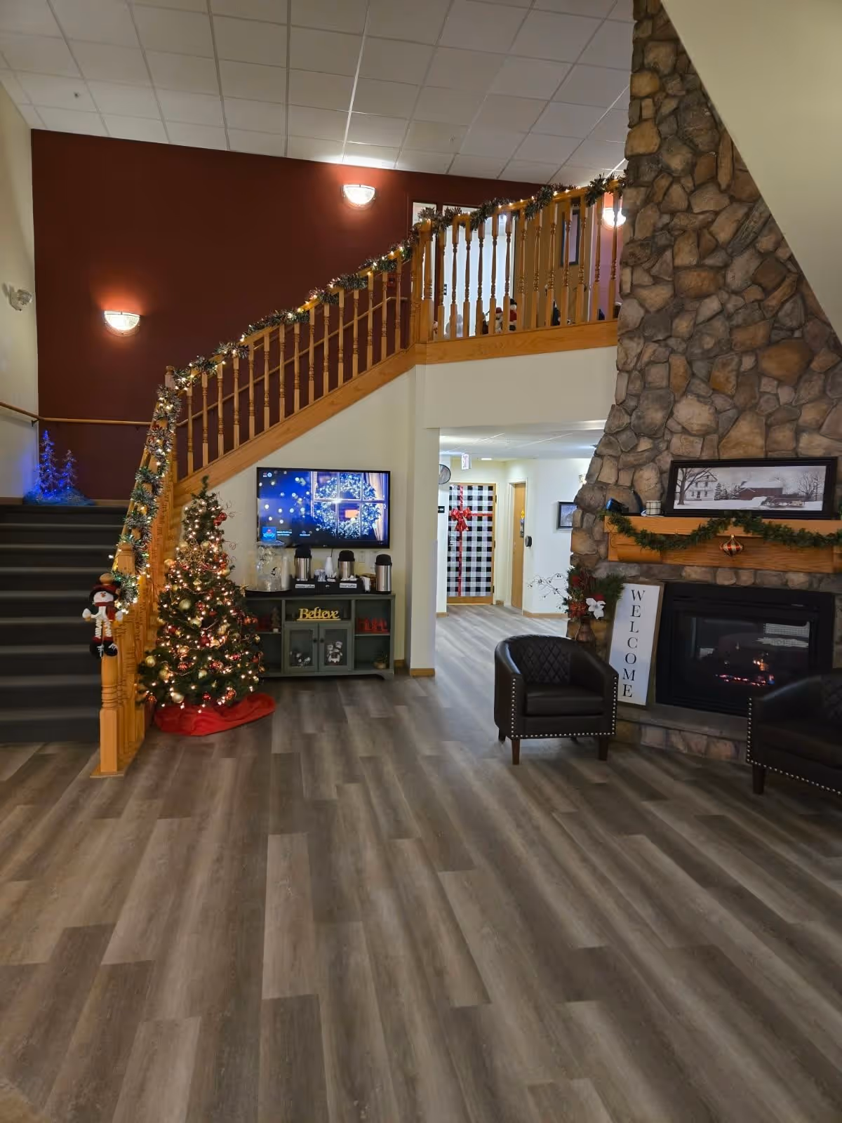 Interior of a senior living facility lobby decorated for Christmas with a small Christmas tree, garlands on the staircase railing, a stone fireplace with a 'Welcome' sign, two black armchairs, and a TV mounted on the wall above a cabinet with coffee supplies.
