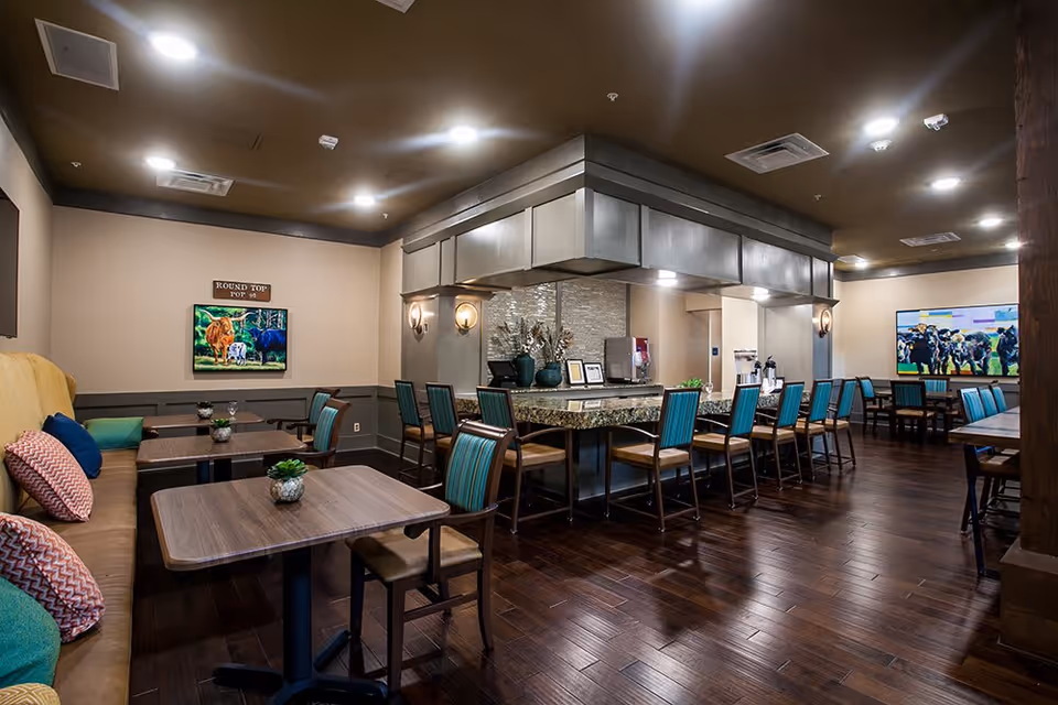 Interior view of a dining area at The Delaney at Lake Waco featuring multiple tables with chairs, a long counter with high chairs, decorative plants, and paintings of cows on the walls. The room has warm lighting and wooden flooring.