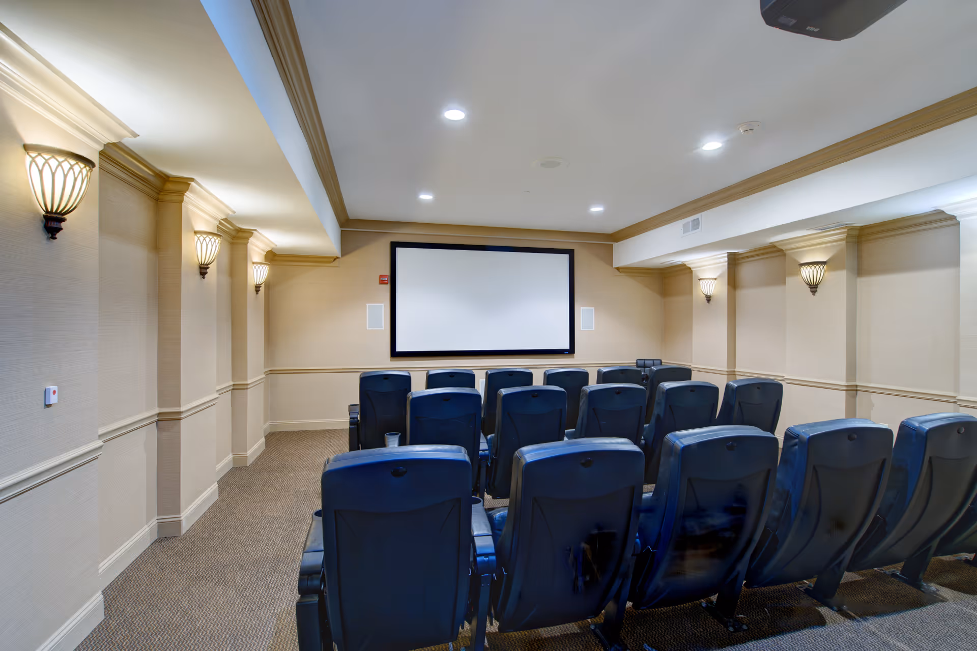 A small theater room with three rows of black cushioned chairs facing a large blank projection screen mounted on a beige wall. The room has beige walls with decorative molding and wall sconces providing soft lighting. The ceiling has recessed lights and a projector mounted near the back.