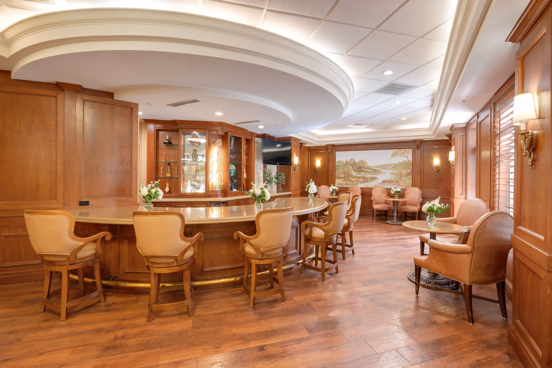 An elegant senior living common area with a curved wooden bar surrounded by leather bar stools. The room features wood-paneled walls, hardwood floors, small round tables with leather chairs, floral centerpieces, wall sconces, and a large landscape painting on the far wall.