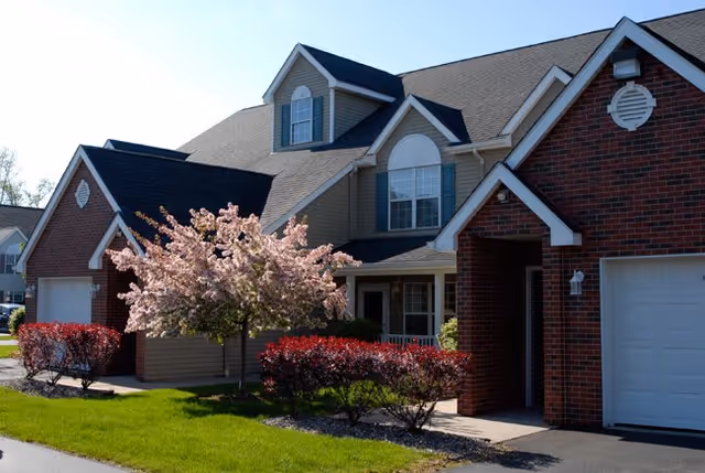 Exterior view of a residential-style senior living facility building with brick and beige siding, featuring a blooming pink tree and neatly trimmed red bushes in front, under a clear blue sky.
