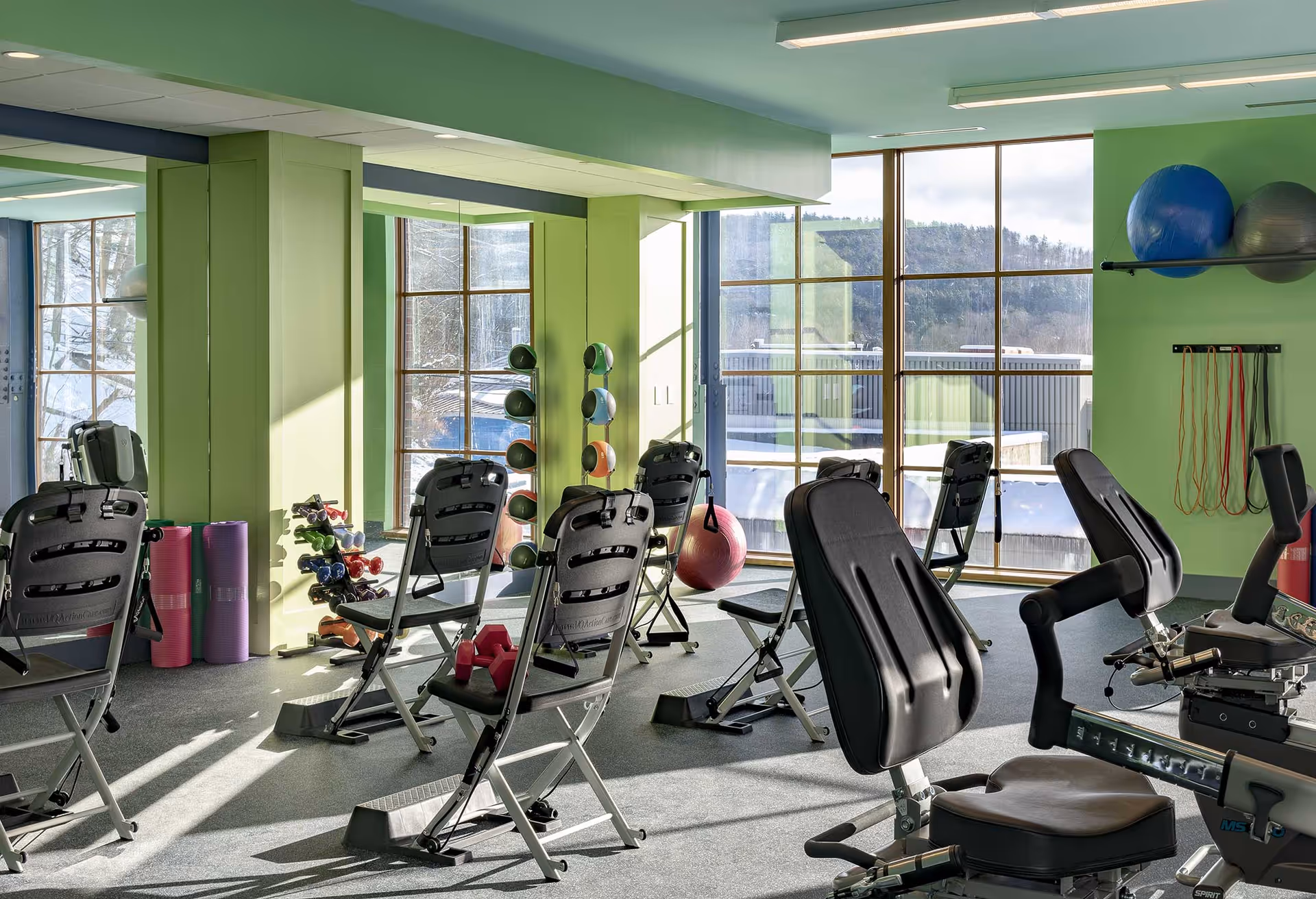 A bright exercise room with large windows showing an outdoor view. The room has green walls and contains several exercise chairs, dumbbells, exercise balls, resistance bands, and yoga mats. Mirrors cover one wall, reflecting the equipment and natural light.
