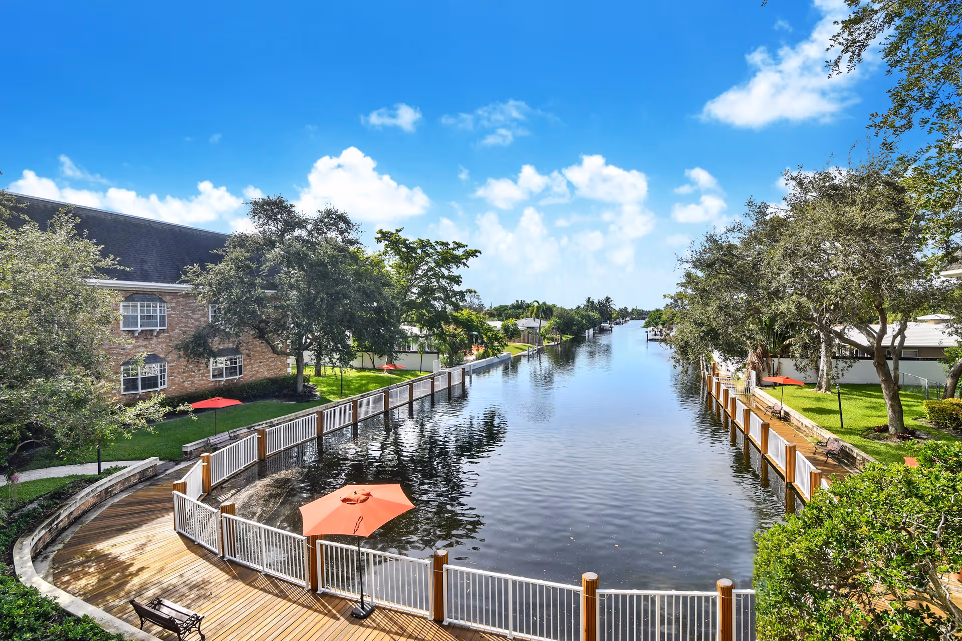A scenic view of a calm water canal bordered by a wooden deck with white railings and orange umbrellas. There are green lawns with trees and benches on both sides of the canal, and a brick building is visible on the left side under a bright blue sky with scattered clouds.
