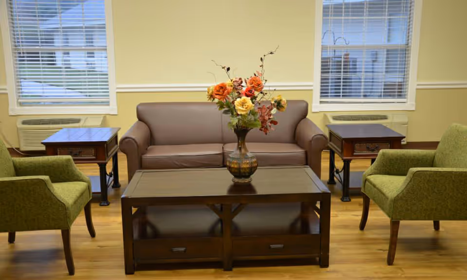 A cozy sitting area with a brown leather sofa, two green upholstered armchairs, a dark wooden coffee table with a vase of colorful flowers, and two matching side tables under windows with white blinds.