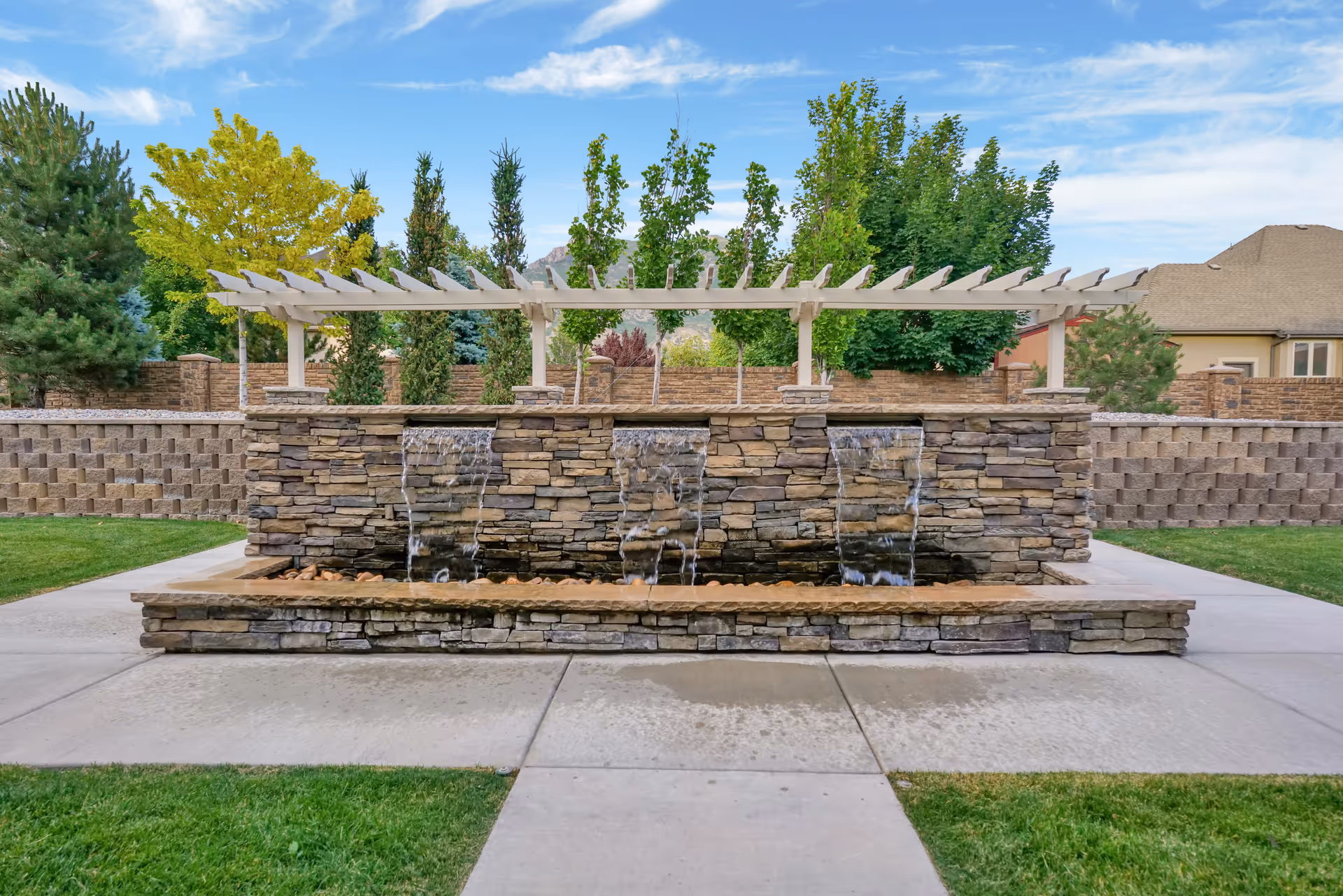 Outdoor stone water feature with three small waterfalls flowing into a rectangular basin, surrounded by a concrete walkway and green grass. Behind the water feature is a white pergola and various trees and shrubs, with a residential building visible in the background under a partly cloudy blue sky.