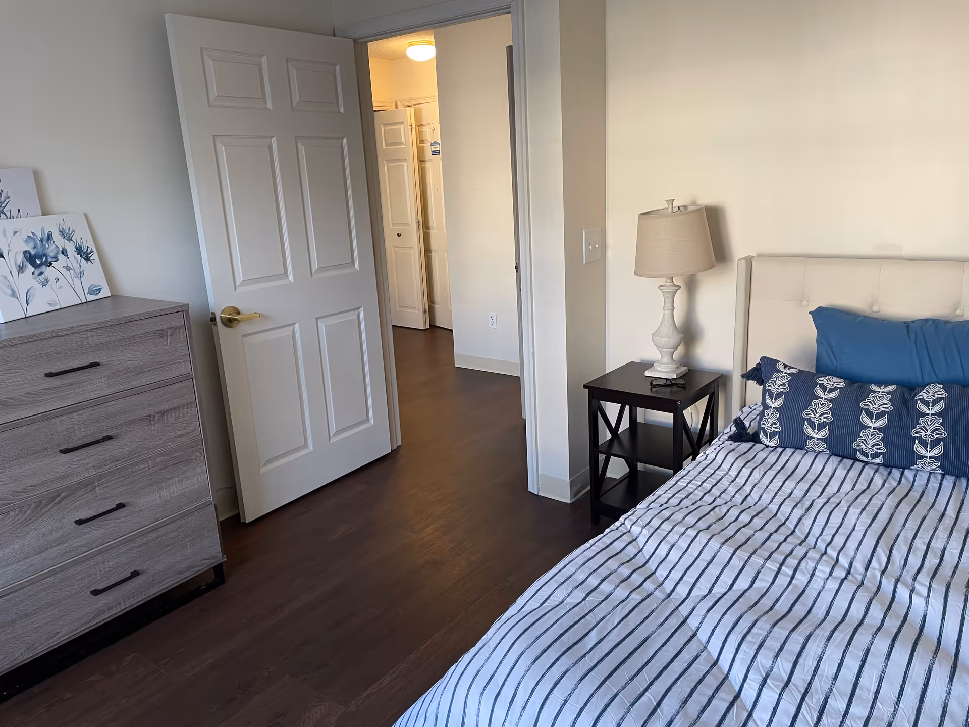 A bedroom with a bed on the right side featuring a white and blue striped bedspread and blue pillows. Next to the bed is a black nightstand with a white lamp. On the left side, there is a wooden dresser with three drawers and two floral paintings resting on top. The door to the room is open, showing a hallway with wooden flooring and white walls.