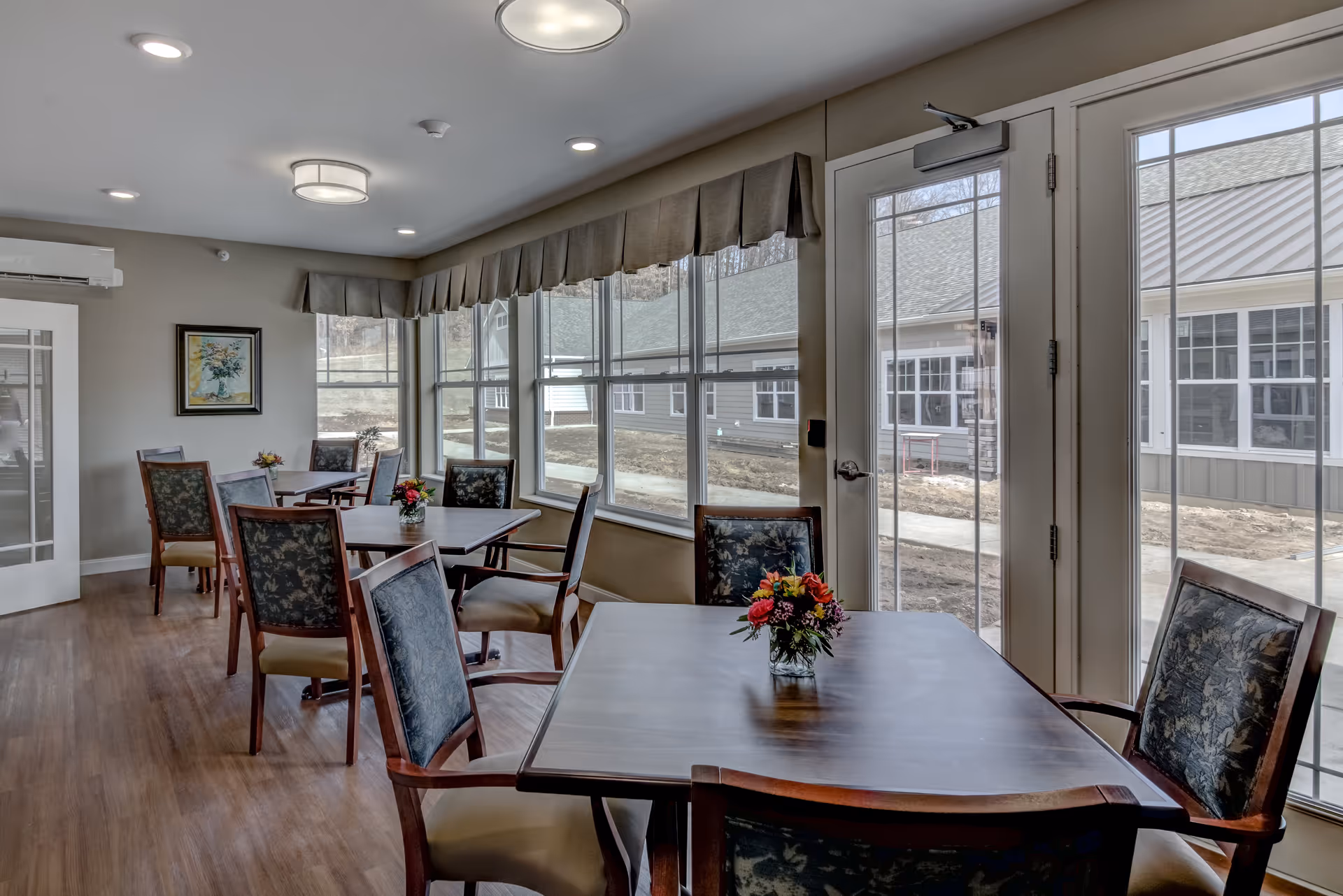 A bright dining room with several wooden tables and chairs arranged neatly. Each table has a small floral centerpiece. Large windows and glass doors let in natural light and offer a view of the outdoor area with buildings and a walkway. The room has light-colored walls, wood flooring, and ceiling lights.