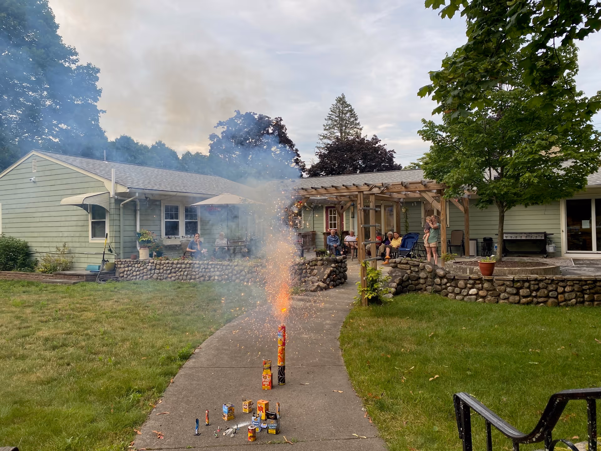 Outdoor scene at Commonwealth House Assisted Living showing a pathway with fireworks igniting and sparking. In the background, several people are seated and standing near a green building with a stone wall and a wooden pergola. Trees and grass surround the area under a cloudy sky.