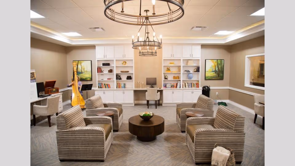 A cozy living room area with four striped armchairs arranged around a round wooden coffee table. Behind the seating area are white built-in shelves filled with books and decorative items, and a desk with a computer and chair. The room features two large circular chandeliers hanging from a tray ceiling. There are two framed landscape paintings on the walls, and a person playing a harp in the background near a record player.