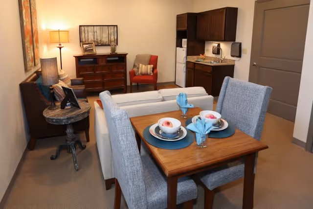 A cozy interior living space featuring a small wooden dining table set for two with blue placemats, napkins, and cups. Behind the table is a light-colored sofa facing a wooden dresser with a framed picture and a lamp. To the right, there is a compact kitchenette with dark cabinets, a sink, a microwave, and a refrigerator. A red armchair with a pillow is positioned near the dresser, and the room is softly lit with warm lighting.