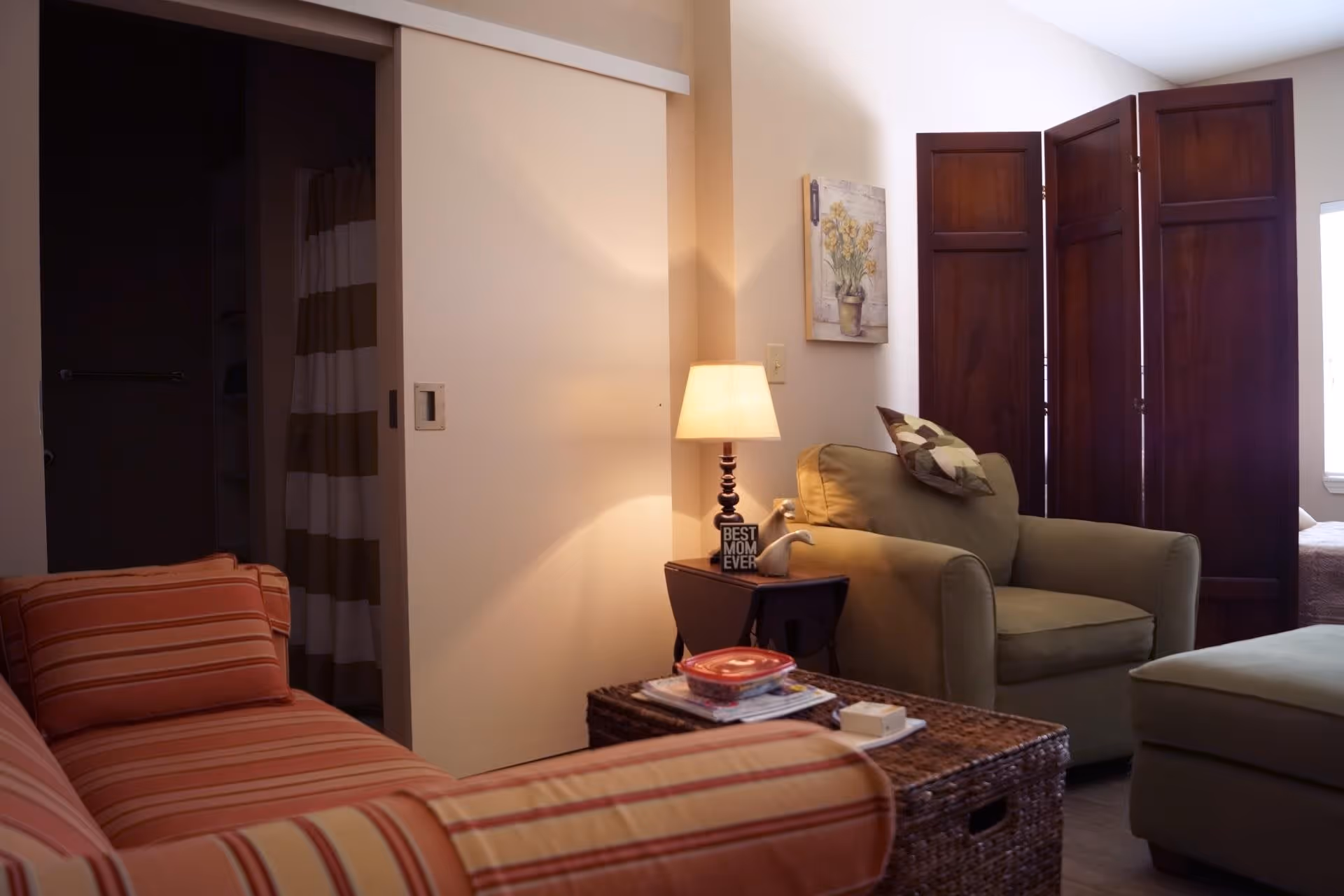 A cozy living room area with a striped orange couch, a green armchair with a patterned pillow, a wicker coffee table with items on it, a side table with a lamp and a decorative sign that reads 'BEST MOM EVER', a folding wooden room divider, and a partially visible bathroom with a striped shower curtain.