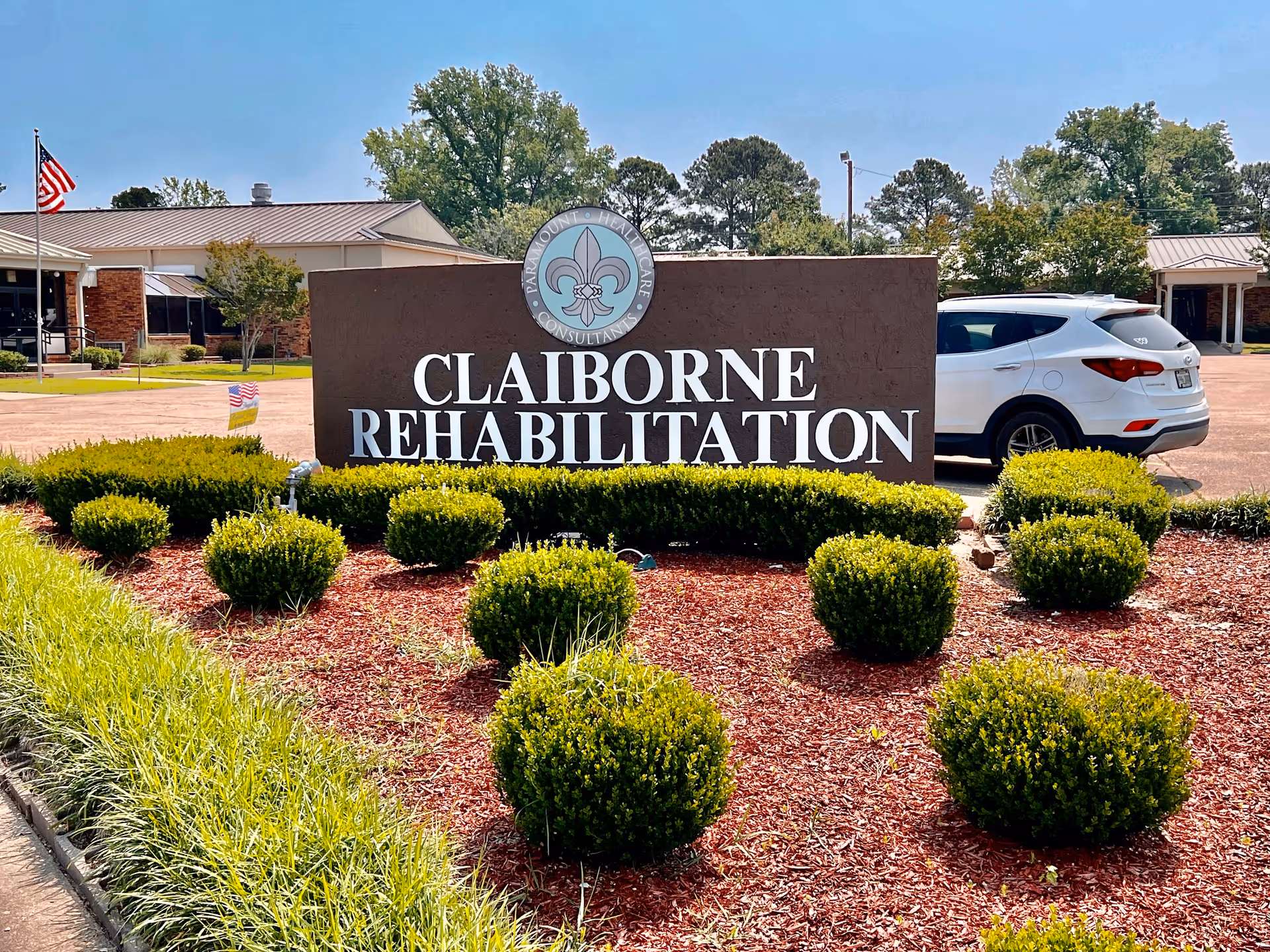 Outdoor view of Claiborne Rehabilitation facility sign surrounded by neatly trimmed bushes and mulch with a white SUV parked nearby and a building and American flag in the background under a clear blue sky.