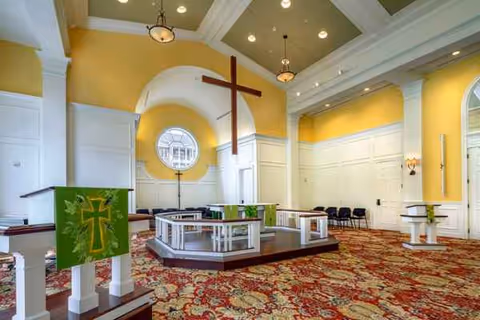 Chapel-like interior with cross, altar, banners, circular window, yellow walls, ornate carpet.
