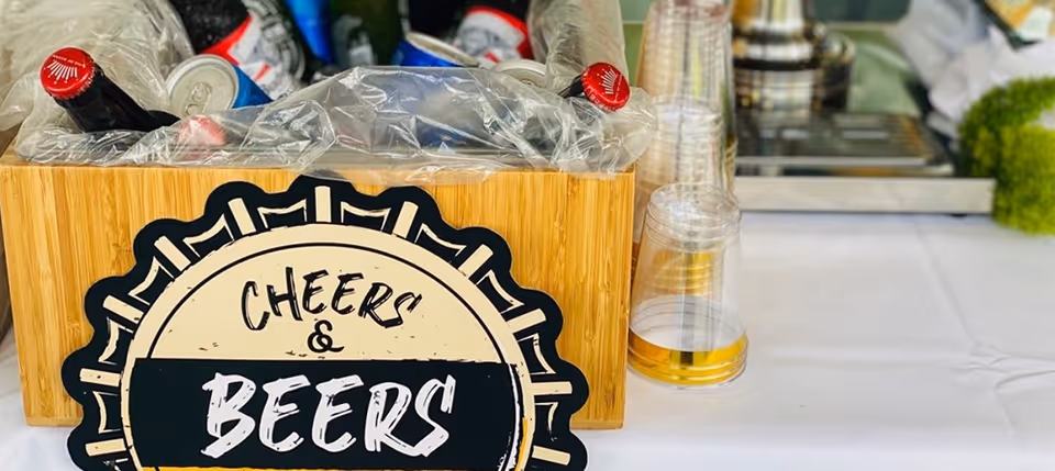 A wooden ice-filled box of bottled beers with a round 'Cheers & Beers' sign and stacked plastic cups on a table.