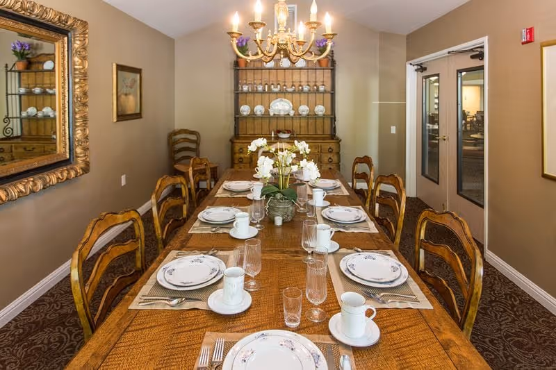 A formal dining room with a long wooden table set for eight people with white plates, cups, and glasses. A floral centerpiece decorates the middle of the table. The room has beige walls, a chandelier hanging above the table, a large mirror on the left wall, and a wooden hutch with decorative plates at the far end. Double doors with glass panels are visible on the right side.