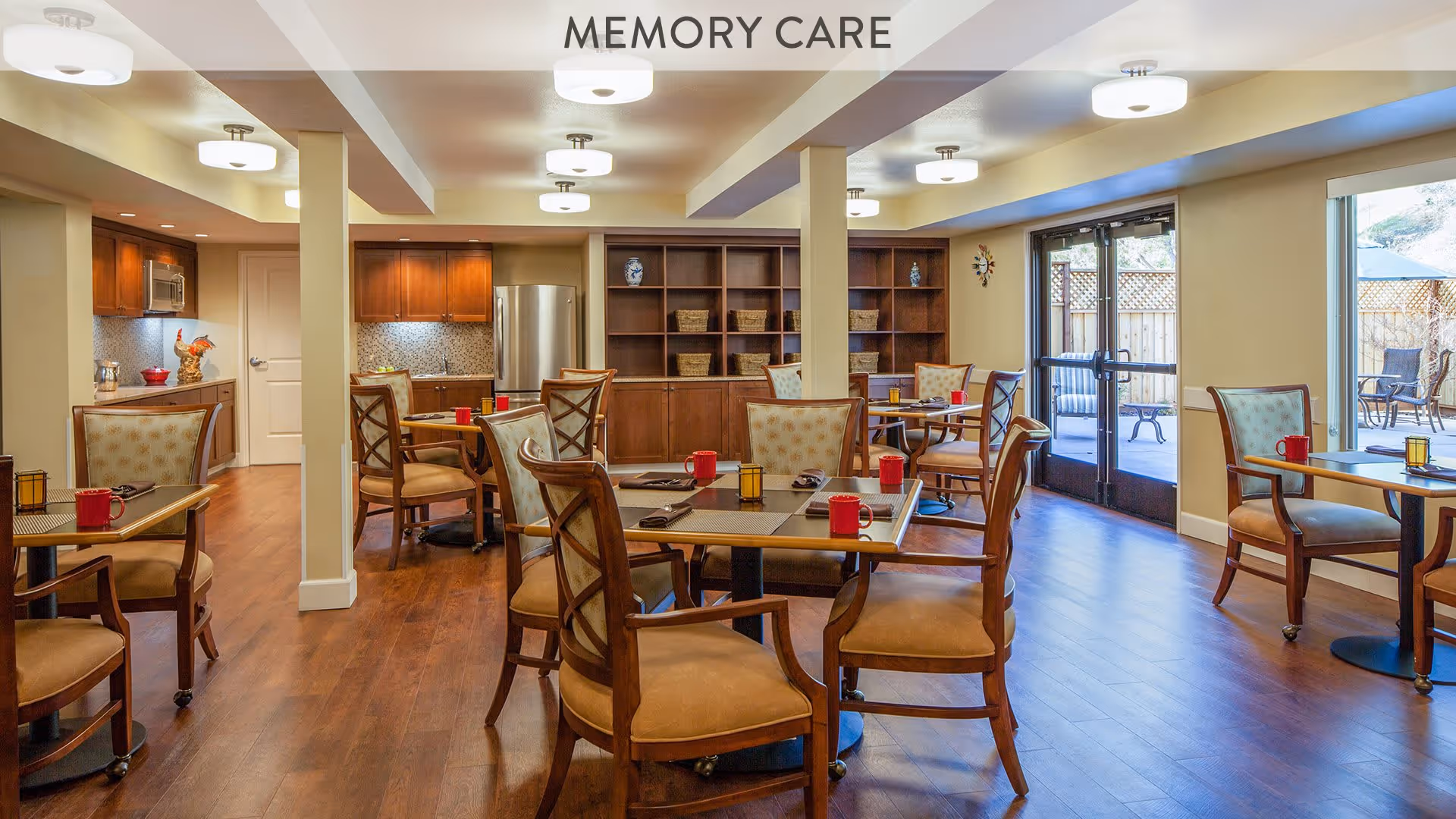 A well-lit memory care dining room with wooden floors, several tables and chairs arranged neatly, red mugs on the tables, a kitchenette area with cabinets and a refrigerator, and large glass doors leading to an outdoor patio area.