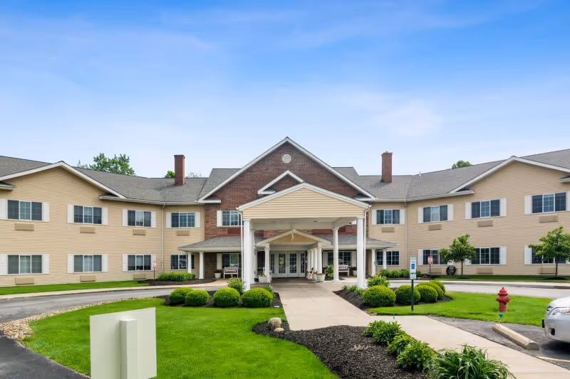 Front exterior view of Eden Vista Stow, a two-story senior living facility with beige siding and a brick central section. The entrance features a covered driveway with white columns, surrounded by well-maintained green lawns, shrubs, and a clear blue sky.