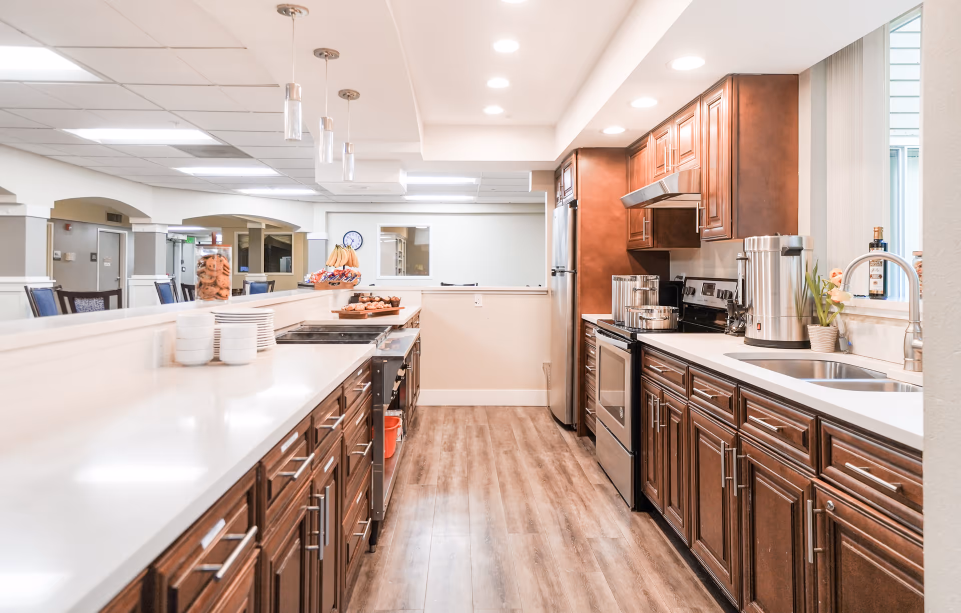 A bright and clean kitchen area with wooden cabinets, stainless steel appliances including a refrigerator and stove, a double sink, and a countertop with various kitchen items such as a large coffee urn, plates, and snacks. The kitchen opens to a dining area with tables and chairs visible in the background.