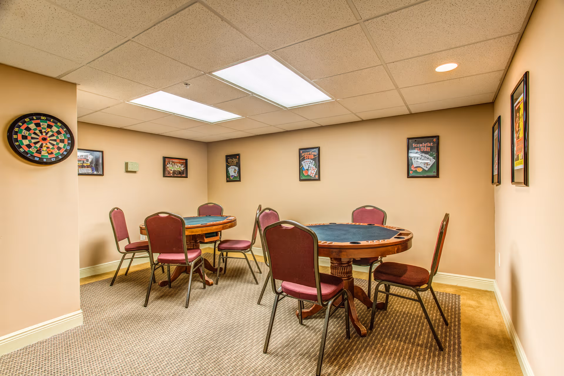 A small game room with two round card tables surrounded by maroon chairs. The walls are beige and decorated with framed pictures related to card games. A dartboard is mounted on the left wall. The room has a carpeted floor and a drop ceiling with fluorescent lighting.
