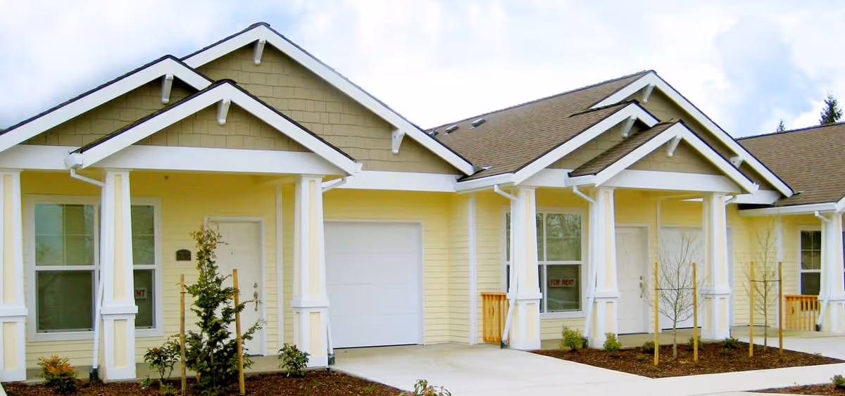 Front exterior of a yellow single-story residential building with covered porches, garage doors, and small landscaped beds.