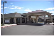 Exterior view of Highland Village of Fallon senior living facility showing a covered entrance with a driveway, beige building with arched windows, and a clear blue sky.