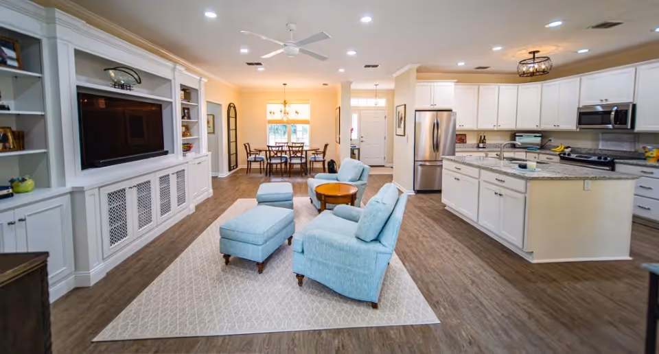 A spacious open-concept living area featuring light blue upholstered chairs and ottomans arranged on a patterned rug in front of a built-in white entertainment center with a large flat-screen TV. The room extends to a dining area with a wooden table and chairs near a window, and a modern kitchen with white cabinetry, granite countertops, stainless steel appliances, and a central island with a sink. The space has wood flooring and neutral-colored walls with ceiling lights and a ceiling fan.