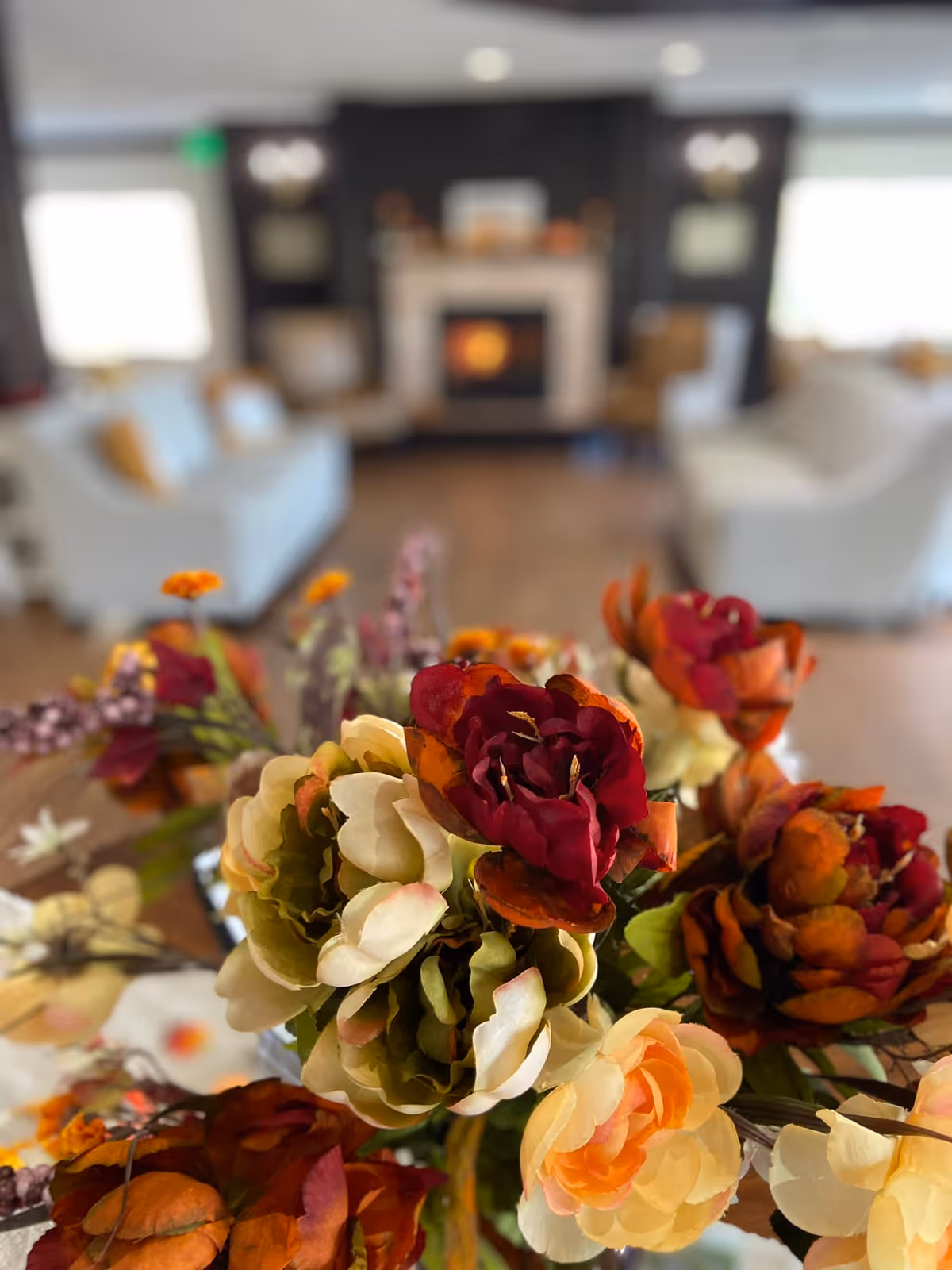 Close-up of a bouquet of colorful artificial flowers in the foreground with a blurred background showing a cozy living room featuring white sofas and a fireplace.