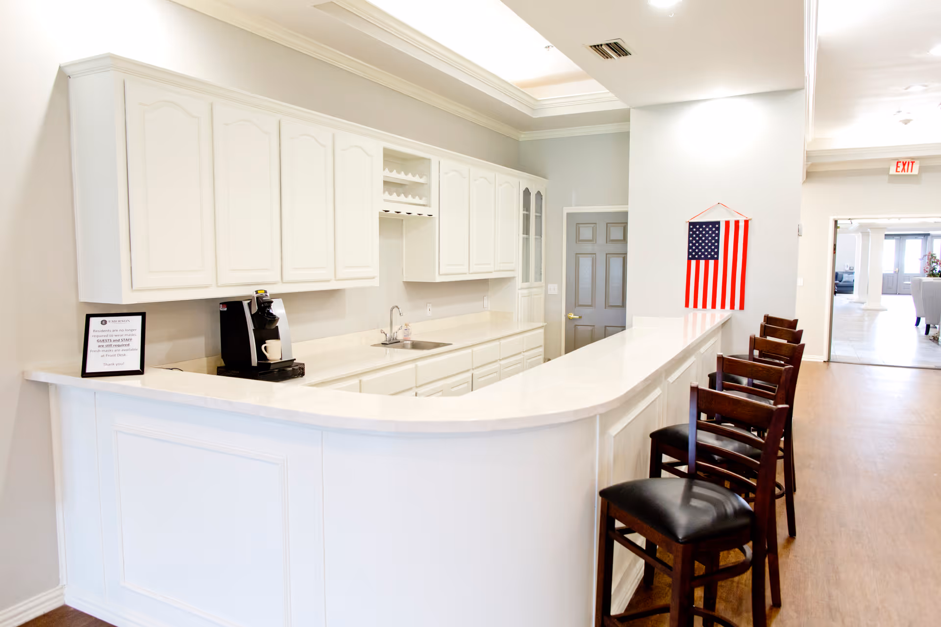 A bright and clean kitchen area with white cabinets and countertops. There is a coffee machine on the counter and a small sink. Four wooden chairs with black cushions are lined up along the curved counter. An American flag hangs on the wall near a closed gray door. The floor is wooden, and the space extends into a hallway with more seating visible in the background.