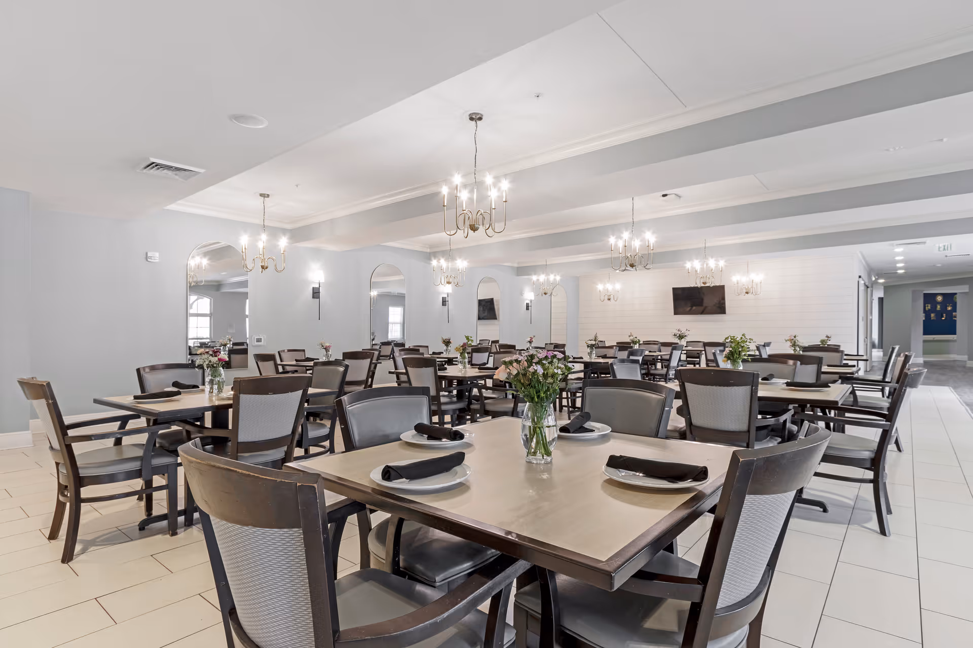 A spacious dining room with multiple wooden tables and chairs arranged neatly. Each table has a small vase with flowers and place settings with plates and black napkins. The room features chandeliers hanging from the ceiling, large mirrors on the walls, and a television mounted on the far wall. The floor is tiled in light colors, and the overall decor is modern and elegant.
