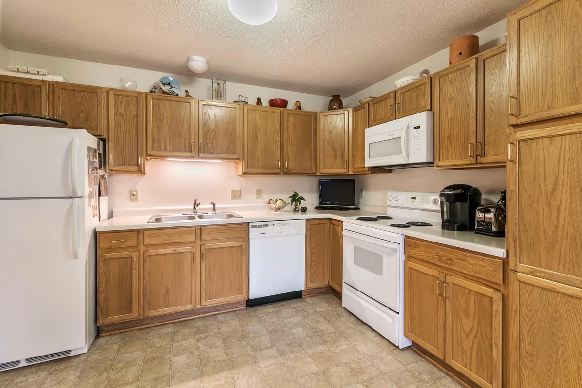 A kitchen with wooden cabinets, a white refrigerator, a white stove with an oven, a white microwave above the stove, a dishwasher, a double sink, and various small appliances including a coffee maker and toaster. The floor is tiled, and there are decorative items on top of the cabinets.