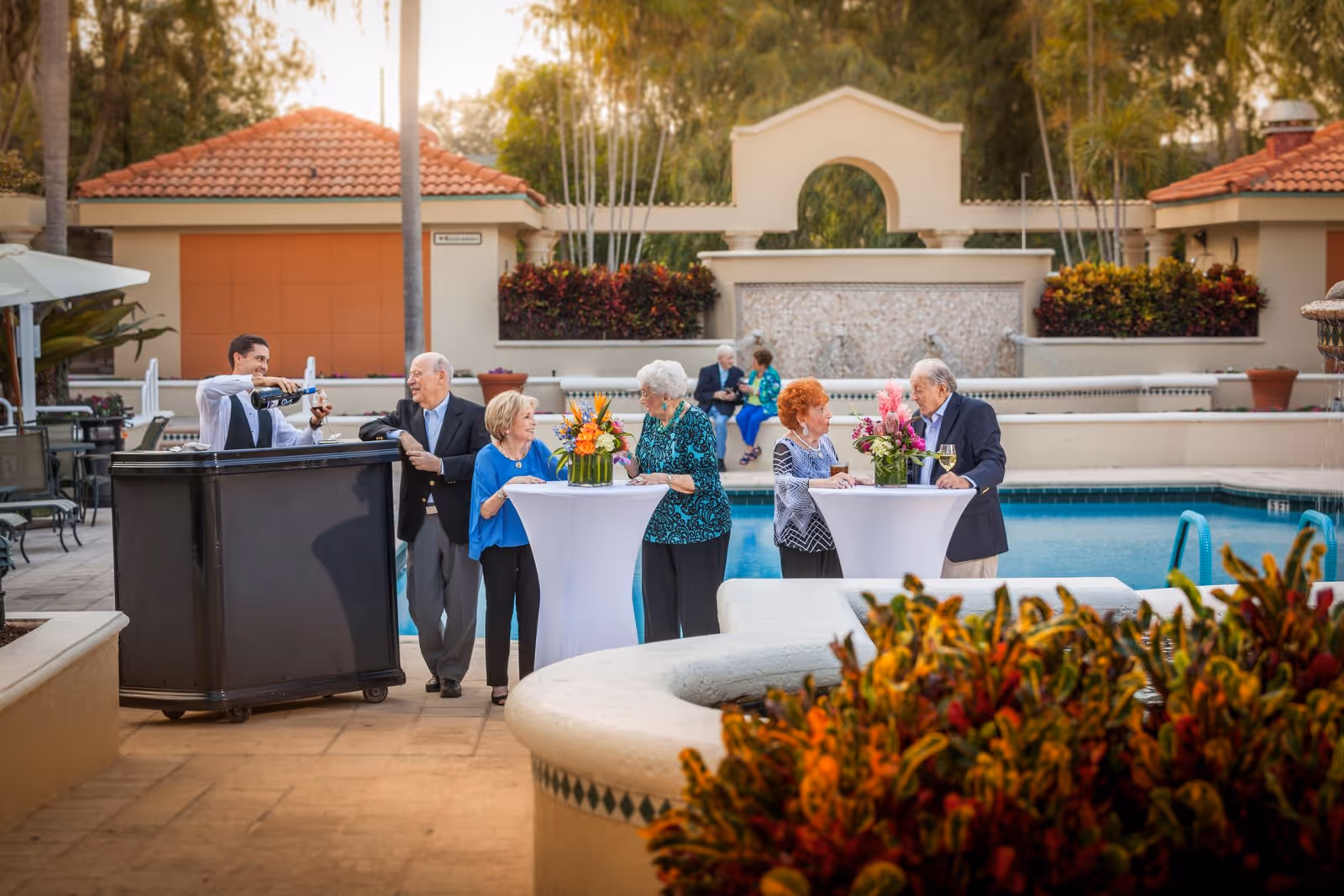 A group of elderly people socializing outdoors near a swimming pool at a senior living facility. Two cocktail tables with floral centerpieces are set up, and a man is pouring wine from behind a portable bar. The setting includes tiled roofs, plants, and a decorative wall with a water feature in the background.
