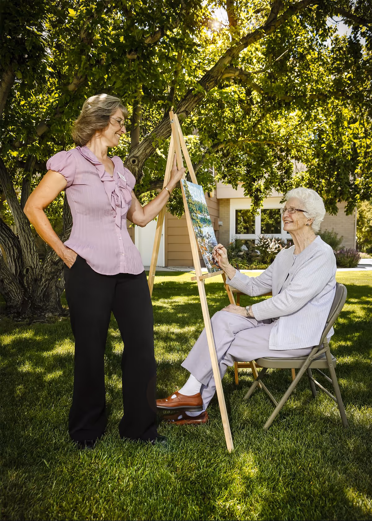 An elderly woman sitting on a folding chair outdoors painting on an easel, while a younger woman stands beside her smiling. They are in a grassy area with trees and a building in the background.