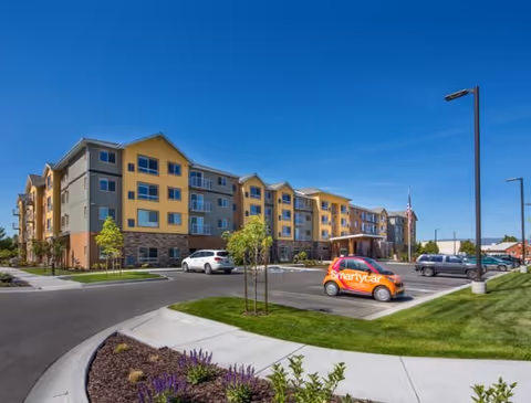 Exterior view of a multi-story senior living facility building with a parking lot in front. The building has a combination of yellow and beige walls with stone accents on the lower level. Several cars are parked, including a small orange car with the word 'Smartycar' on its side. The sky is clear and blue, and there are landscaped areas with grass, small trees, and flowers around the parking lot.