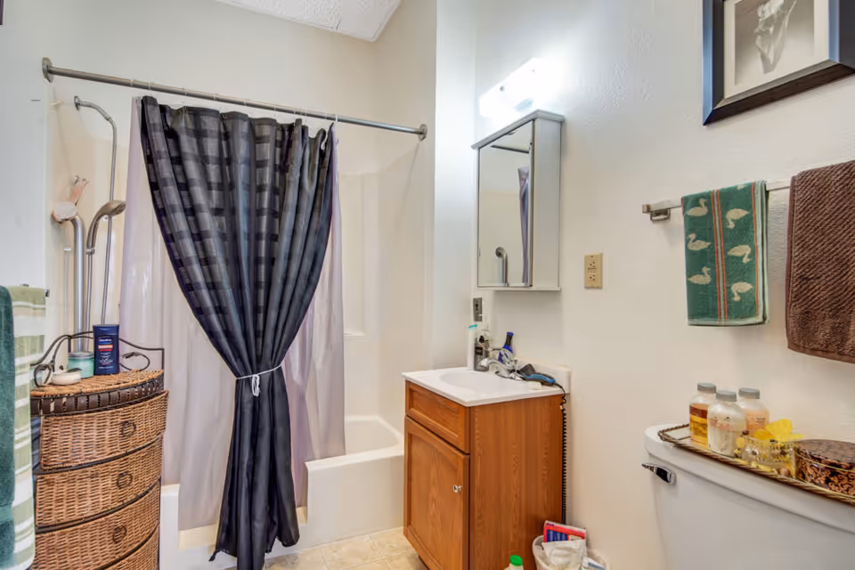 A bathroom with a bathtub and shower combination featuring a black and gray checkered shower curtain tied in the middle. There is a wicker storage unit with toiletries on top next to the tub. A wooden vanity with a sink and a mirrored medicine cabinet above it is on the right side. Two towels, one green with a duck pattern and one brown, hang on a towel rack above the toilet, which has a tray with bottles and a decorative box on the tank. The walls are light-colored and there is a framed picture above the towels.