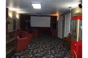 A dimly lit room with patterned carpet, red upholstered chairs, a popcorn machine on the right, and a large white projection screen on the far wall, resembling a small theater or media room.