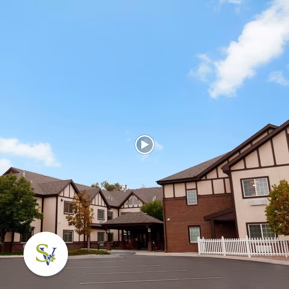 Two-story Tudor-style assisted living building entrance with a parking lot, white picket fence, and trees under a clear blue sky.