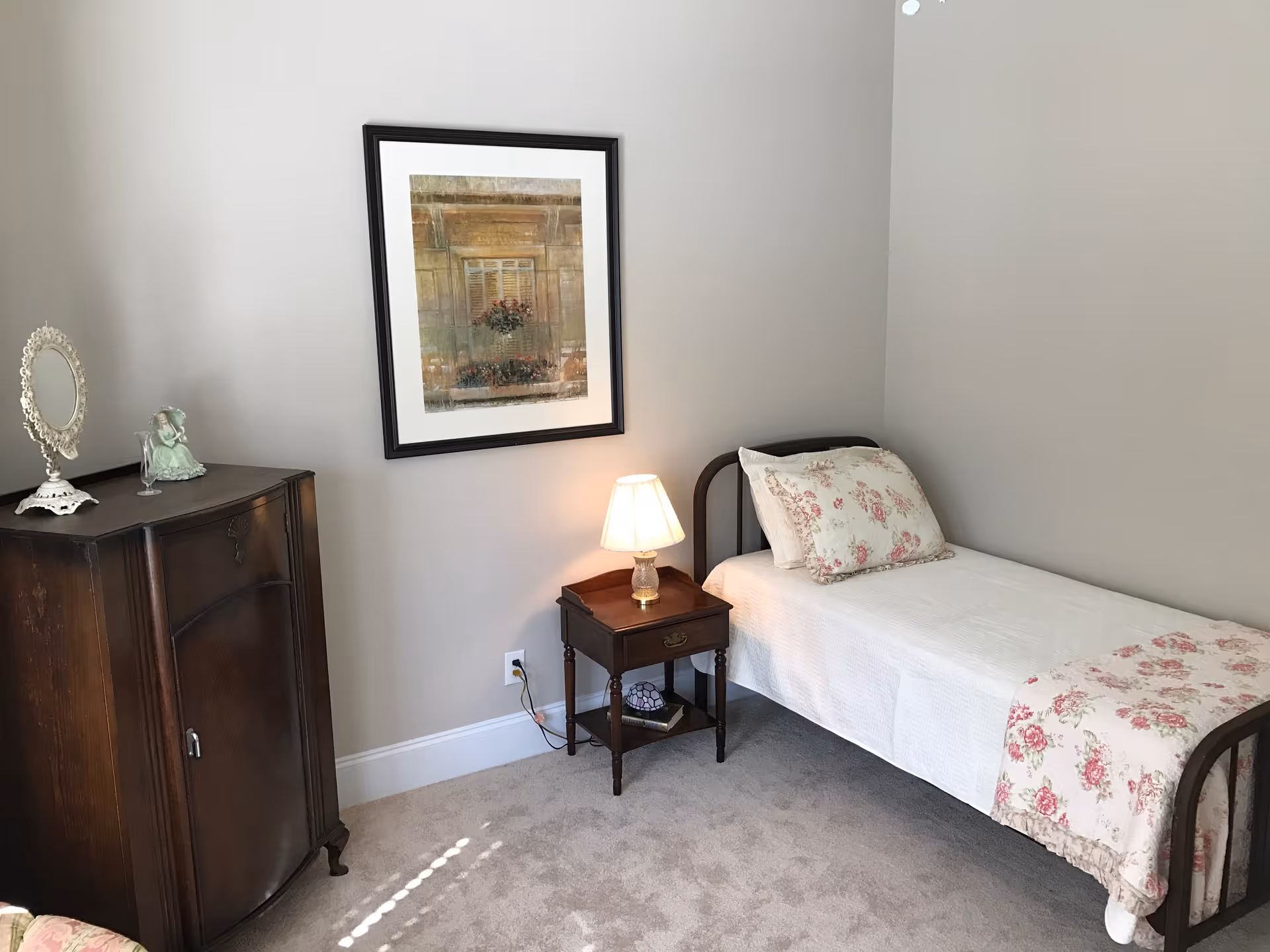 A small bedroom with a single bed covered in white bedding and floral pillows and a matching throw. Next to the bed is a wooden nightstand with a lit table lamp and a book on the lower shelf. On the left side of the room is a dark wooden cabinet with decorative items on top, including a small mirror and a figurine. A framed picture hangs on the light gray wall above the nightstand. The floor is carpeted in a light beige color.