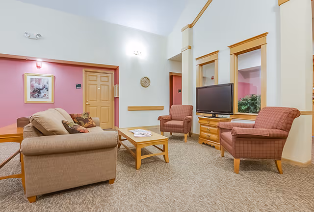 A cozy living room area in a senior living facility with a beige sofa, two red armchairs, a wooden coffee table with magazines, a wooden TV stand with a flat-screen TV, and a wall clock. The walls are painted in light colors with some pink accents and there is a framed floral picture on the wall near a wooden door.