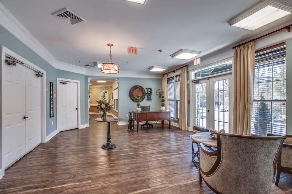 Bright reception lounge with seating, a central table with flowers, a desk against the far wall, and large windows and doors letting in natural light.