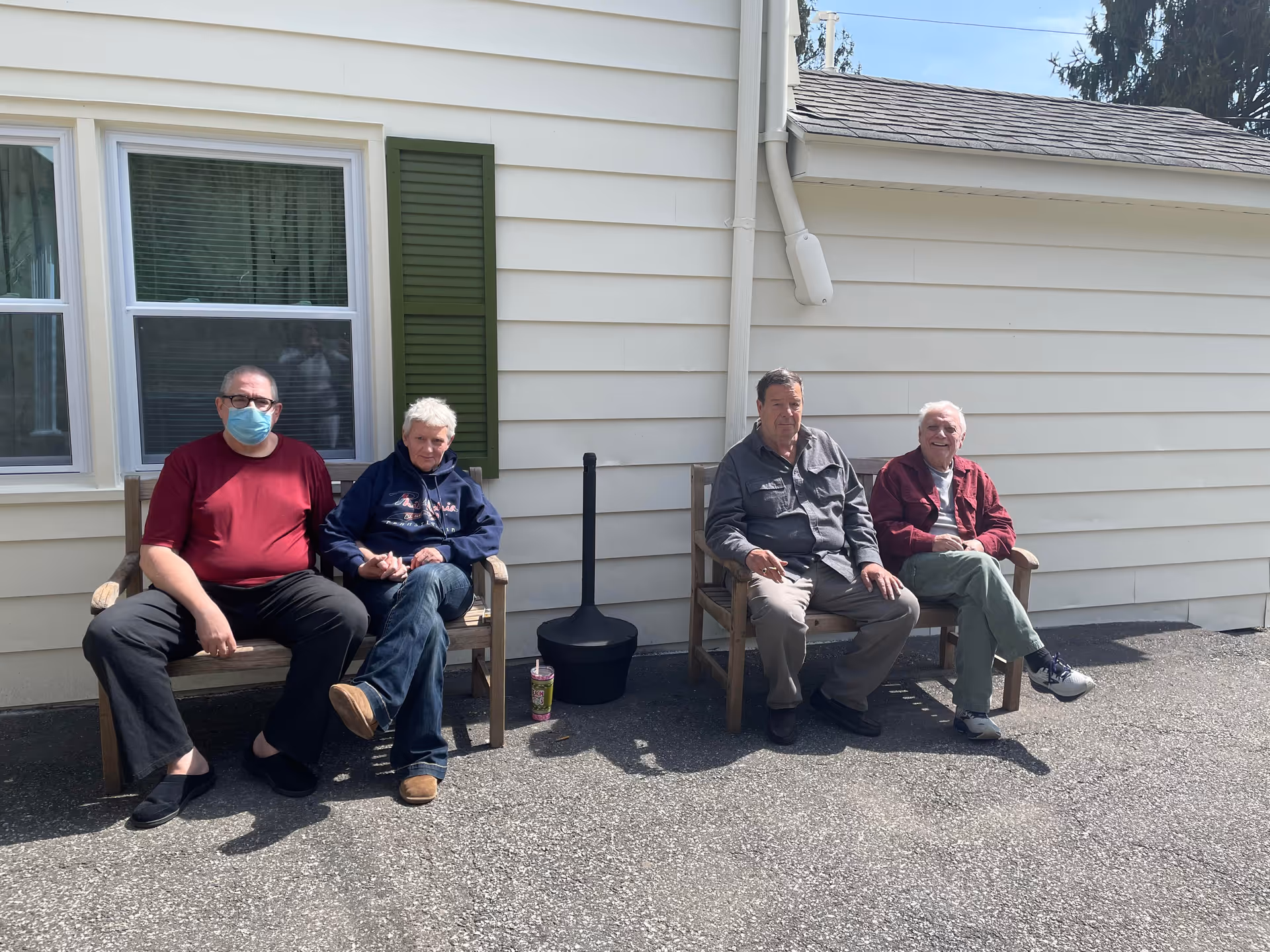 Four elderly men sitting outside on wooden benches against the exterior wall of a building. One man is wearing a red shirt and a face mask, the others are dressed casually. There is a window with green shutters behind them and a small black ashtray or outdoor heater between the benches.