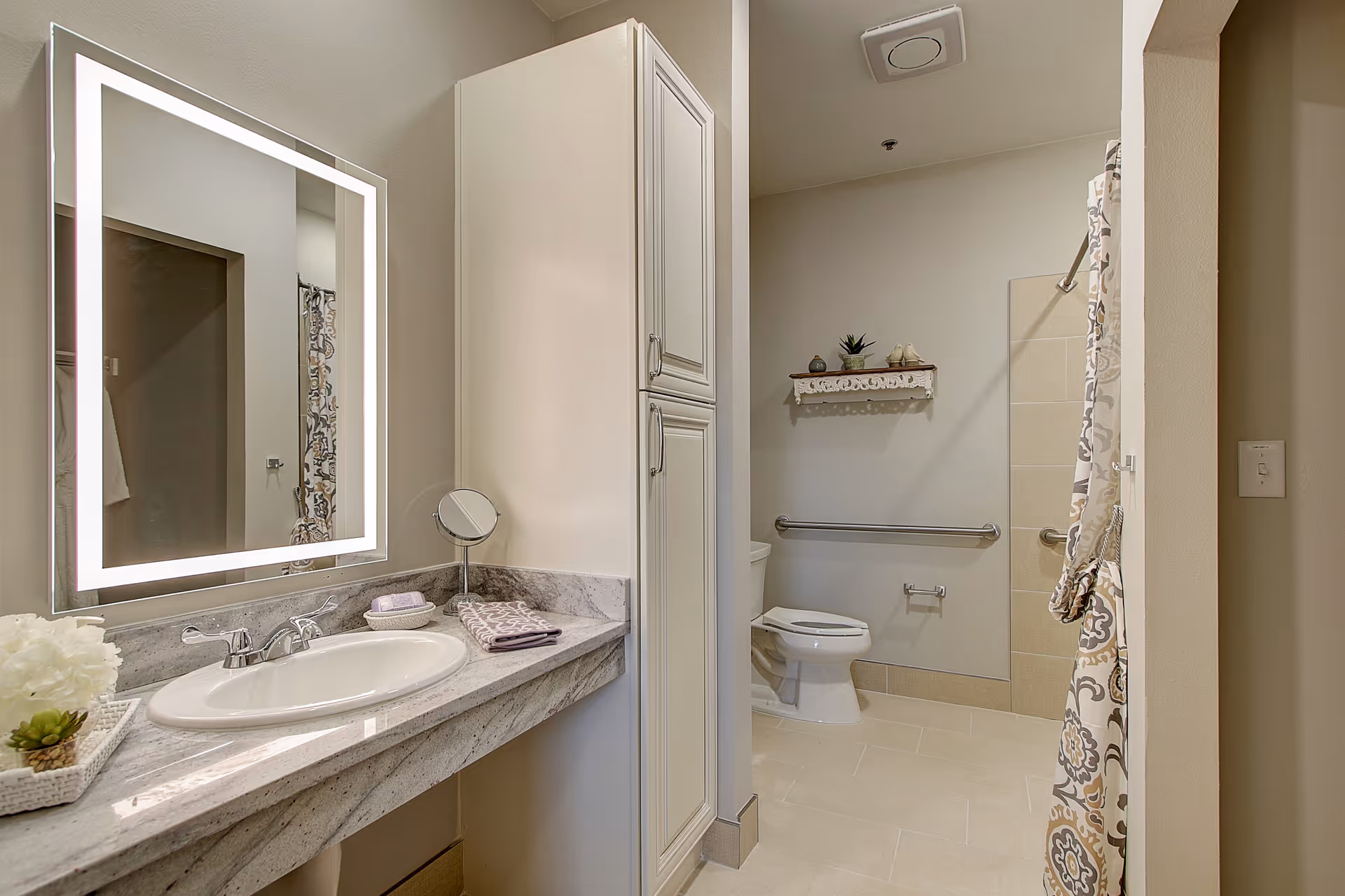 A clean and modern bathroom featuring a large illuminated mirror above a marble countertop with a sink. There is a small round makeup mirror, folded towels, and decorative plants on the counter. The bathroom includes a toilet with grab bars on the wall and a shower area with a patterned curtain tied to the side. A small decorative shelf with plants and ornaments is mounted above the toilet.