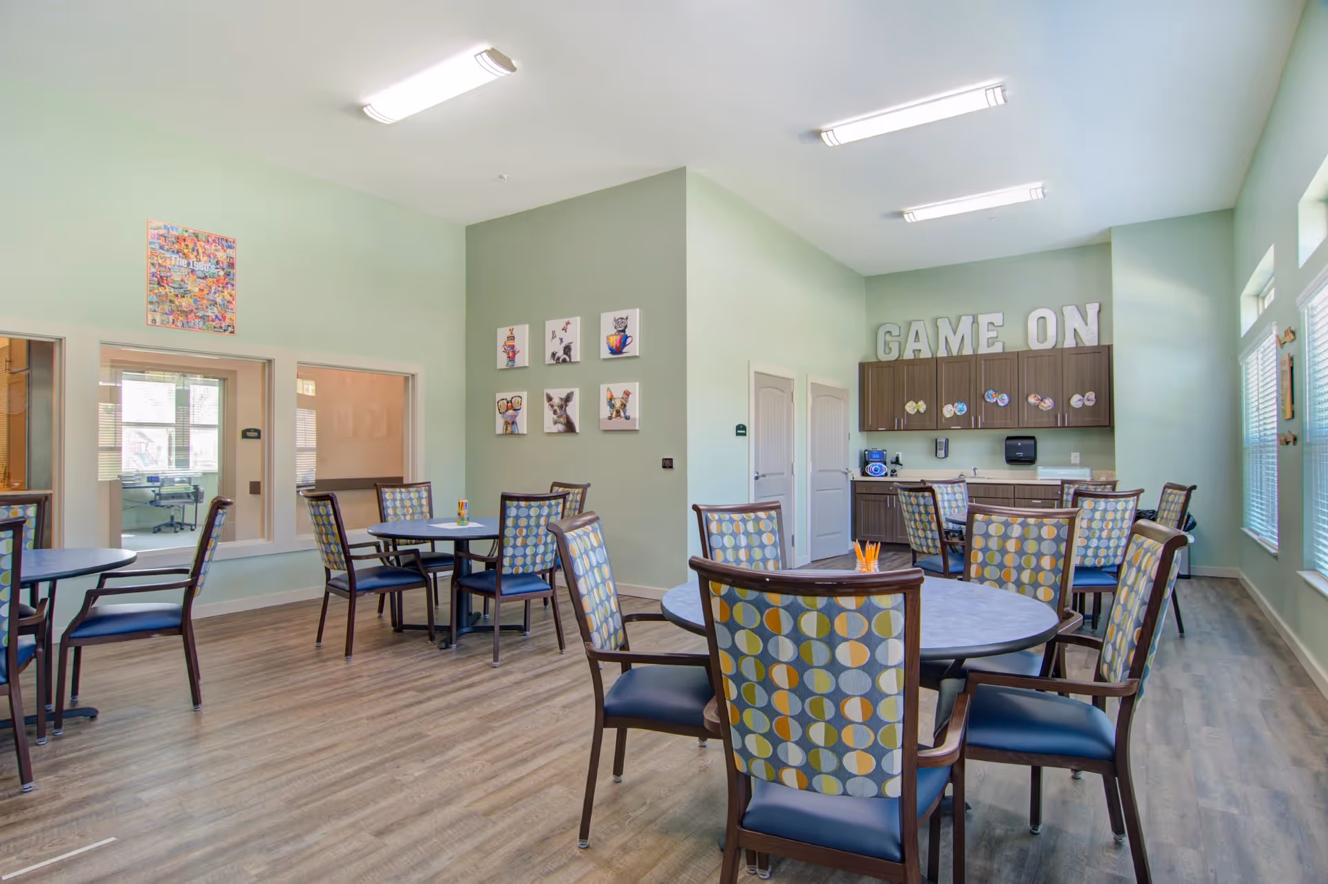 Bright communal activity room with round tables and patterned chairs, a kitchenette area with 'GAME ON' letters on the back wall, and light green walls.