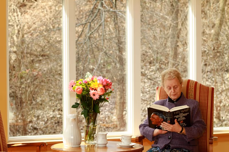 An elderly woman sitting in a striped armchair by large windows, reading a book titled 'His Way'. A round wooden table in front of her holds a vase with colorful flowers, a white pitcher, a small creamer, and a teacup with a saucer. Outside the windows, leafless trees are visible.
