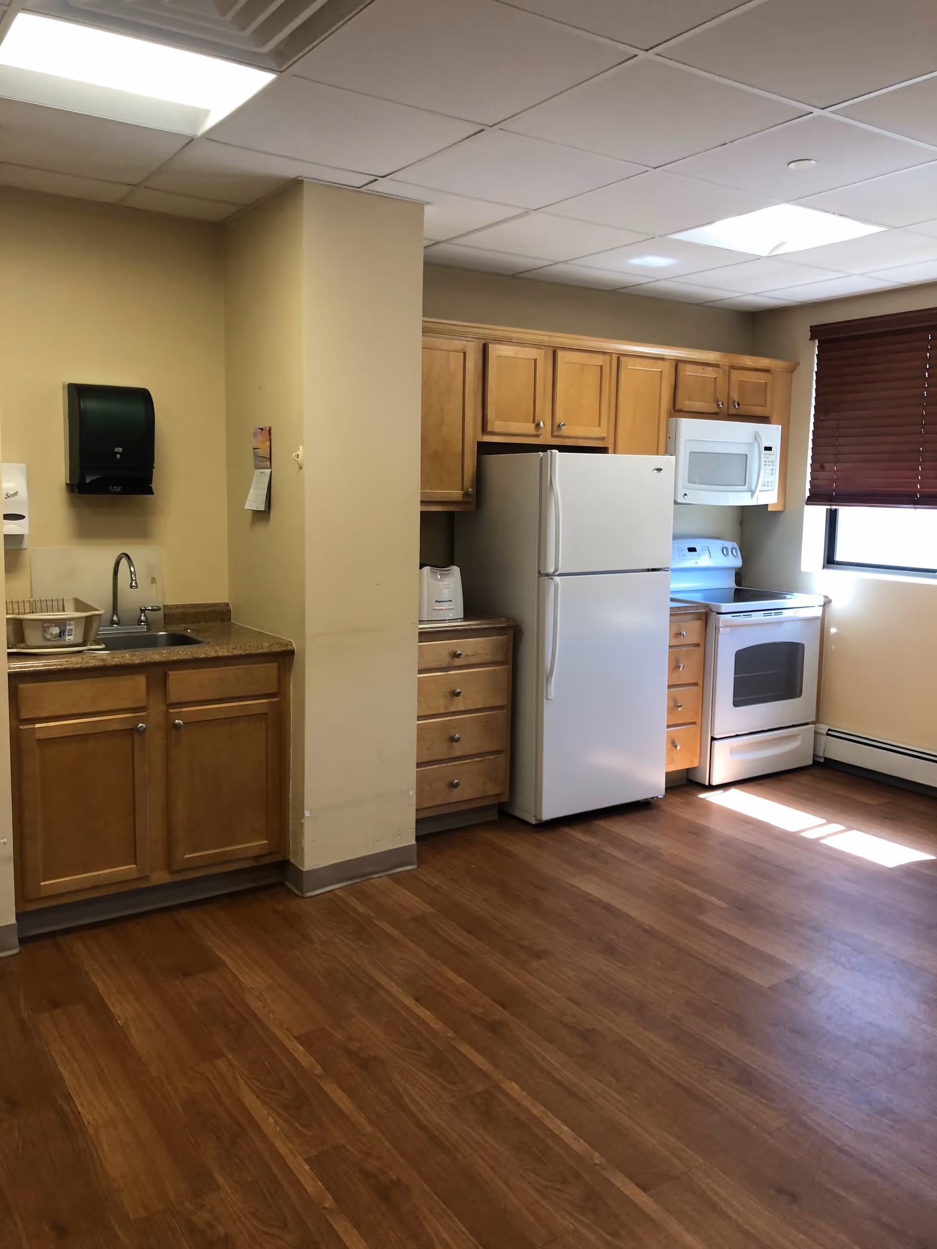 Interior view of a kitchen area with wooden cabinets, a white refrigerator, white microwave, white stove, a sink with a paper towel dispenser above it, and a window with brown blinds letting in sunlight. The floor is wooden and the walls are painted beige.