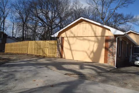 Exterior view of a building with a large closed garage door painted in beige, brick walls on the sides, a wooden fence to the left, and leafless trees in the background under a clear blue sky.