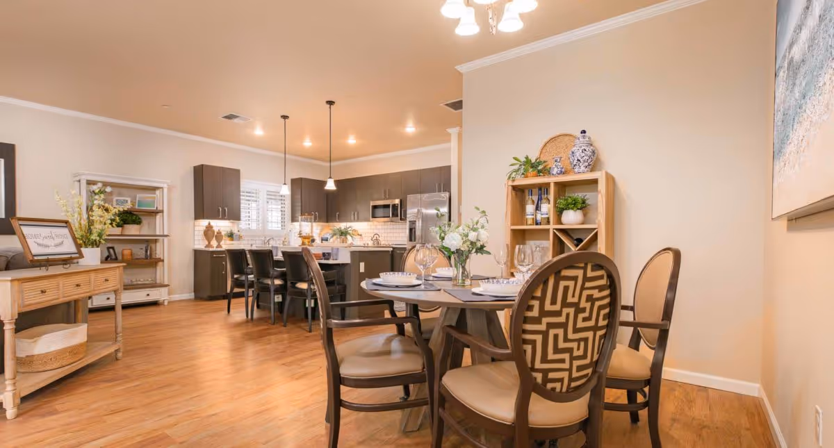 A bright and spacious open-concept dining and kitchen area with wooden flooring. The dining area features a round table set with plates, glasses, and a floral centerpiece, surrounded by four chairs with patterned backs. The kitchen has dark cabinets, a white subway tile backsplash, stainless steel appliances, and a kitchen island with four bar stools. Decorative plants and vases are placed on shelves and countertops, creating a warm and inviting atmosphere.
