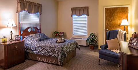 A cozy bedroom in an assisted living community featuring a single bed with patterned bedding, a wooden nightstand with a lamp and framed photos, two windows with valance curtains, a blue armchair with a pillow and blanket, a wooden dresser with framed photos, a potted plant, and a closed door with a lamp beside it.