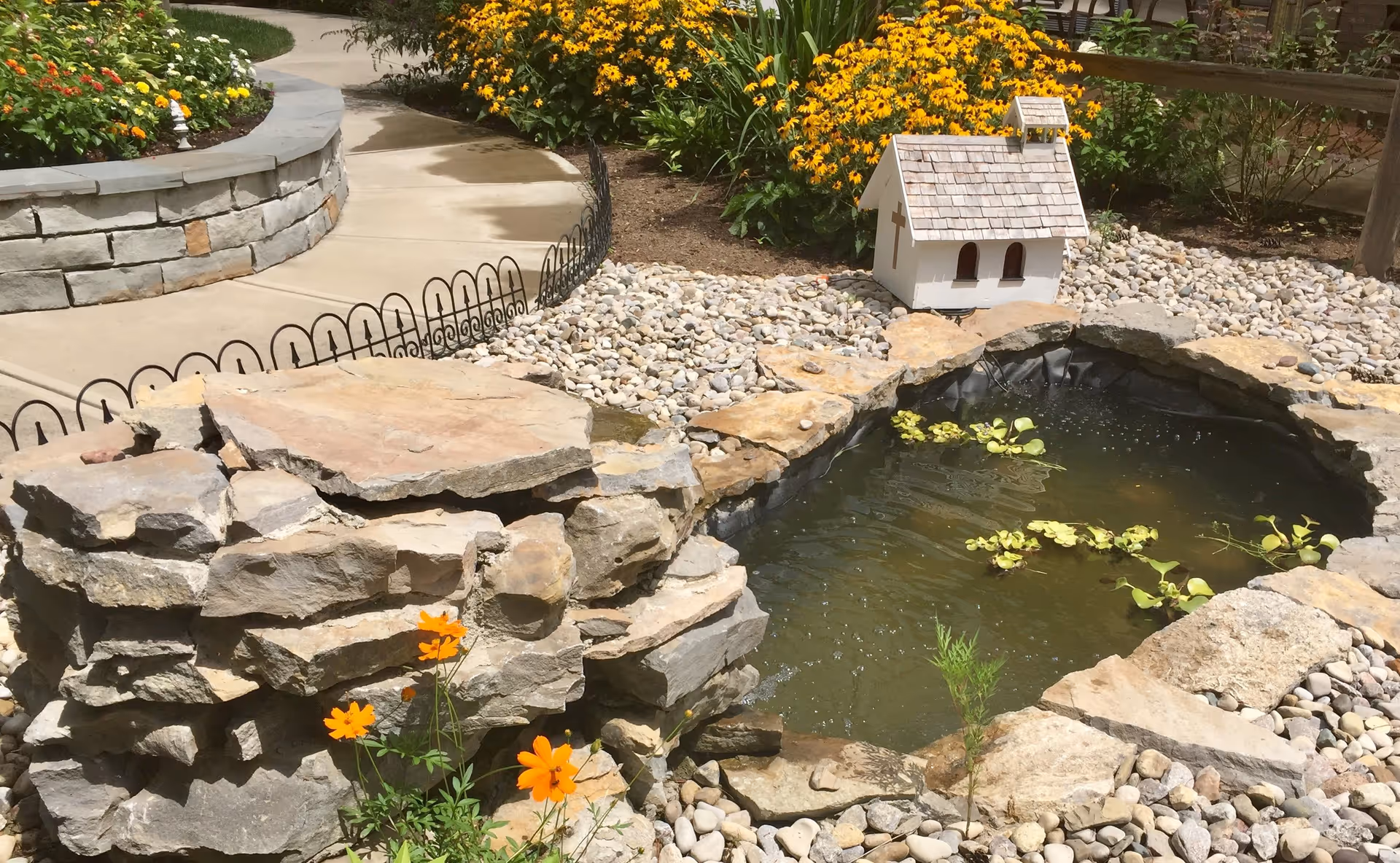 A small outdoor garden area featuring a stone-lined pond with water plants, surrounded by decorative rocks and flowers. A miniature white wooden house with a shingled roof and cross detail is placed near the pond. A curved concrete pathway and a low stone wall with colorful flowers are visible in the background.
